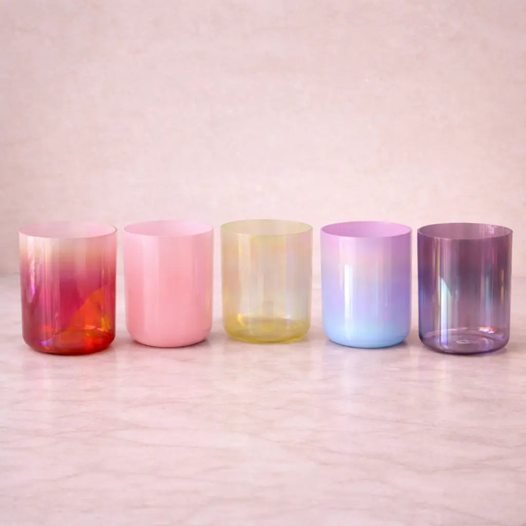 Five iridescent glass tumblers on a wooden surface with a soft pink background