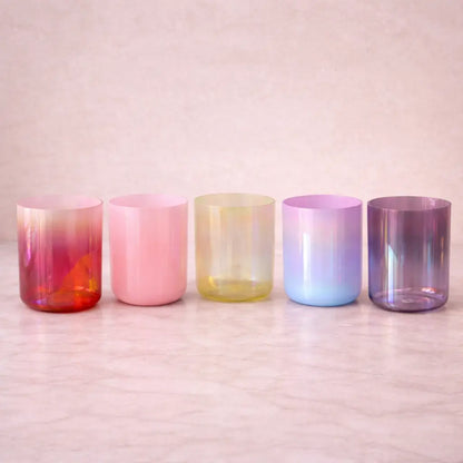 Five iridescent glass tumblers on a wooden surface with a soft pink background